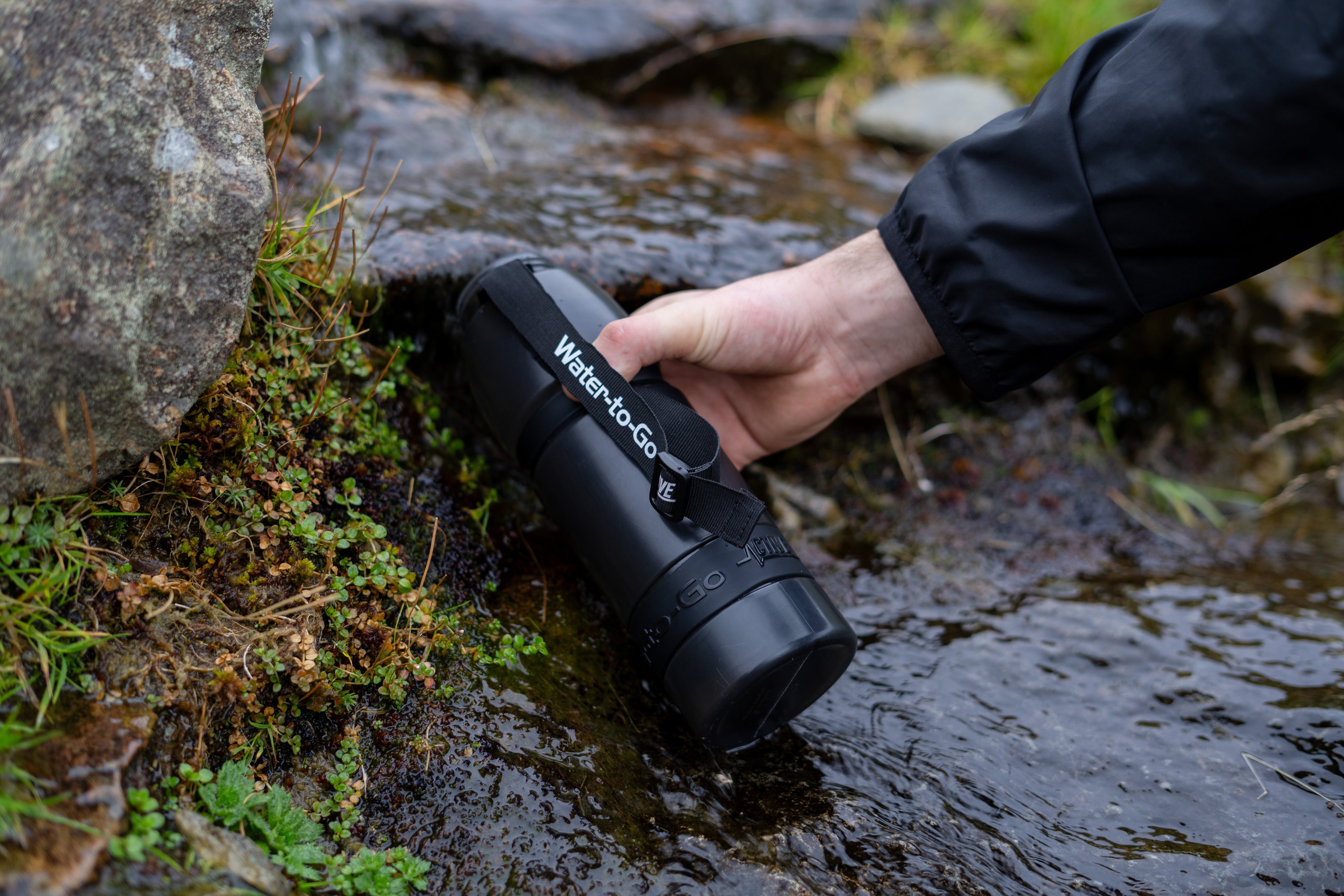 Hand holding a black water filter device labeled 'Water-to-Go' near a stream.