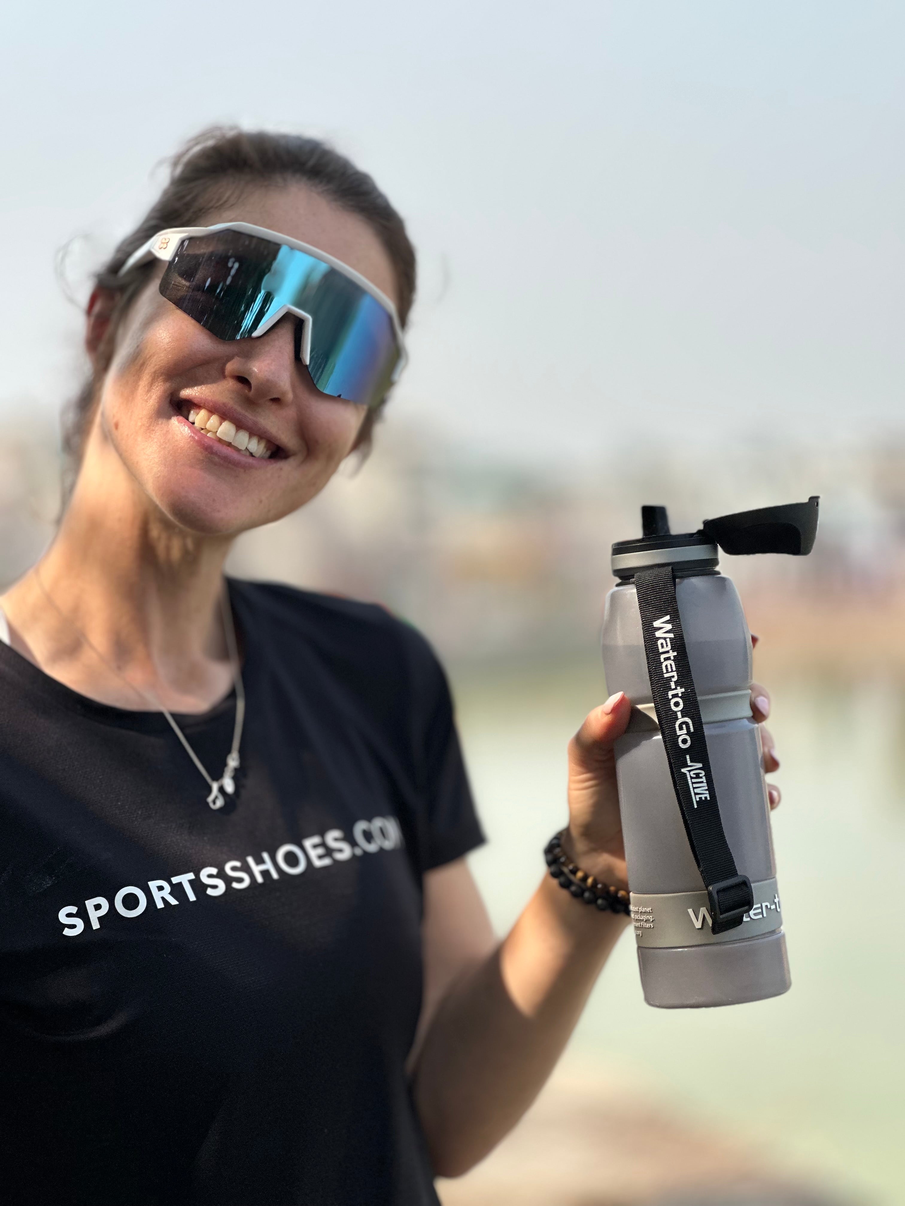 female runner wearing sunglasses, smiling, holding her water to go 1 litre bottle in cloud